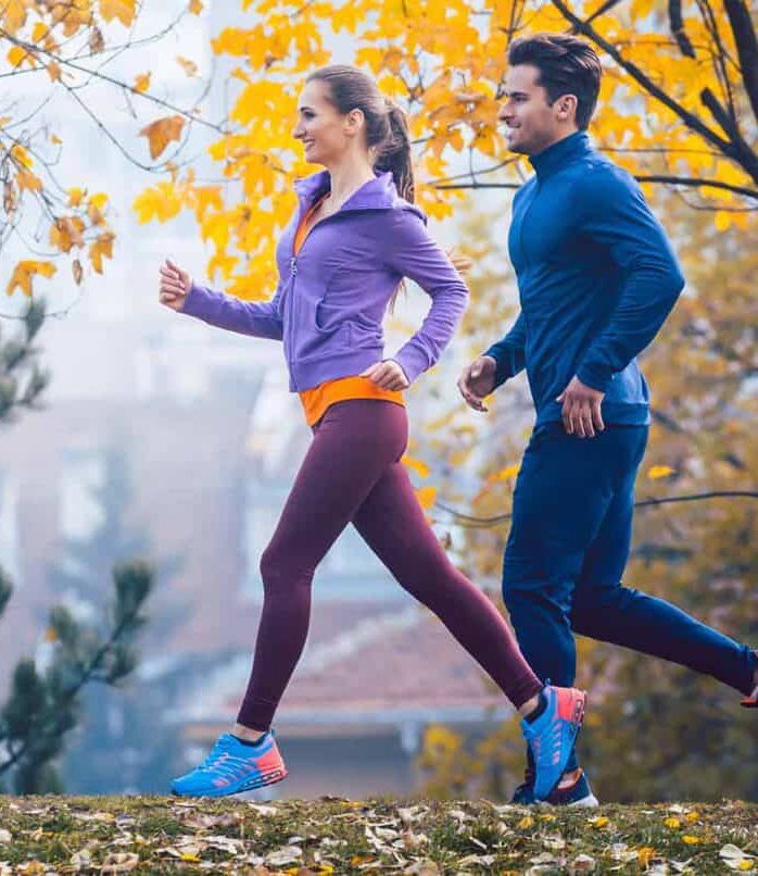 Two people jogging outdoors on a pathway surrounded by autumn leaves. The woman is wearing a purple jacket and pink leggings, while the man is wearing a blue jacket and dark pants.